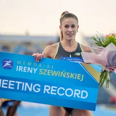 Uśmiechnięta lekkoatletka z bukietem kwiatów trzyma tablicę "MEETING RECORD" z logo memoriału Ireny Szewińskiej.