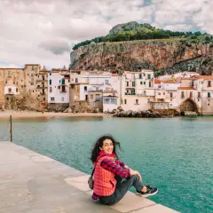 A woman smiles, enjoying her first time visiting Sicily. She sits on a stone pier overlooking the turquoise sea and a picturesque town with a dramatic cliff behind it.