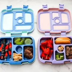 Two bento boxes, one blue and one purple, are filled with lunch items. The blue box contains fruit skewers, cucumber slices, crackers, and ground meat. The purple box holds strawberry skewers, a bun, nuts, and cucumber slices.