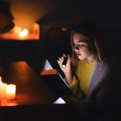 Dziewczyna czyta książkę przy świecach i latarce podczas blackoutu. Co to blackout? To nagłe przerwy w dostawie prądu.