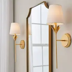 Apliques de pared dorados con pantalla blanca, junto a un espejo con marco dorado.