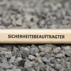 Ein Holzstab mit der Aufschrift "SICHERHEITSBEAUFTRAGTER KITA" liegt auf grauem Schotter.