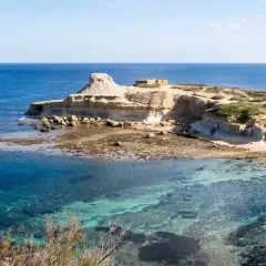 Felsige Küste mit kristallklarem Wasser und kleinen Gebäuden auf der Insel Gozo.