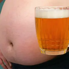 Ein Bierbauch ist da, weil Bier dick macht. Ein Mann hält ein Glas Bier vor seinem Bauch.
