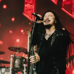 Wokalista zespołu rockowego na scenie, z mikrofonem w dłoni, w tle czerwone światła i perkusja. Koncert w Hali Stulecia we Wrocławiu.