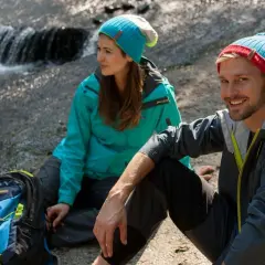 Para, co to jest trekking? Para w czapkach siedzi przy strumieniu, obok plecak.