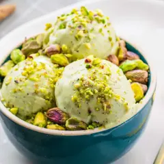 Tre palline di cremoso gelato al pistacchio, guarnite con granella e pistacchi interi, in una ciotola blu.