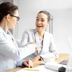 Dwie kobiety w garniturach uśmiechają się i ściskają dłonie przy biurku.