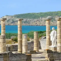 Ruinas romanas en el área de Bolonia, con columnas de piedra y una estatua blanca frente al mar azul y una colina verde.