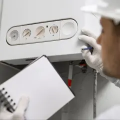 Technik w białej rękawiczce sprawdza ustawienia kotła gazowego kondensacyjnego, aby zapewnić jego optymalne działanie. Notuje dane w zeszycie.