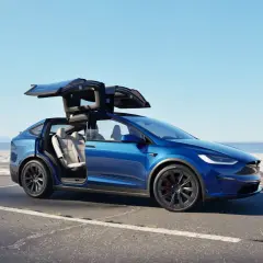 Niebieski Tesla Model X z otwartymi drzwiami typu falcon wing na tle plaży. Sprawdź aktualną tesla model x cena.