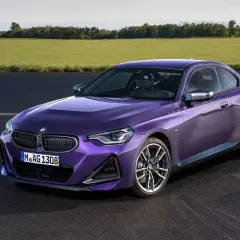 Fioletowe BMW G42 z ciemnymi felgami stoi na asfalcie, za nim zielone pole i drzewa.