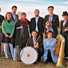 Zespół muzyczny w strojach nawiązujących do stylu folk, z instrumentami: skrzypce, akordeon, bęben, trąbka, klarnet i tuba.