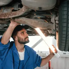 Mechanik w niebieskim kombinezonie oświetla latarką podwozie samochodu, sprawdzając jego stan. Dowiaduje się, ile trwa przegląd samochodu.