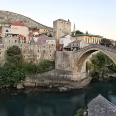 Kamienny, łukowy Mostar most nad turkusową rzeką, otoczony zabytkową zabudową i zielenią.