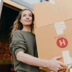 Uśmiechnięta dziewczyna z kręconymi włosami niesie dwa kartony. Na jednym z nich napis "TOYS". To idealny moment na vinted wysyłka za granicę.
