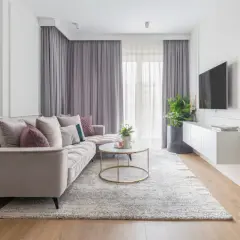 Elegancki salon w stylu klasycznym. Jasna sofa, okrągły stolik kawowy, dywan i zasłony tworzą przytulną atmosferę.