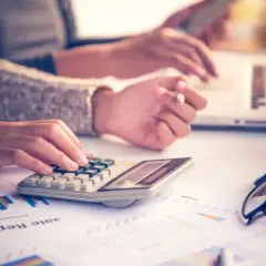 Kalkulacja kosztów podróży służbowej. Dwie osoby pracują przy biurku, jedna używa kalkulatora, druga laptopa.