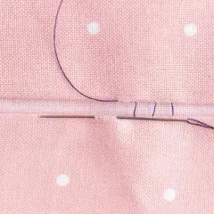 Close-up of a needle and purple thread creating an invisible hem stitch on pink polka-dot fabric.