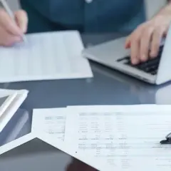 Osoba analizuje dokumenty, porównując umowę o pracę a umowę zlecenie, korzystając z laptopa i kalkulatora.
