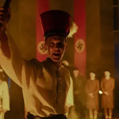 Młody mężczyzna w czerwonym nakryciu głowy z piórem wznosi płonący kij. W tle widać flagi ze swastykami, sugerujące scenę z filmu "Biała odwaga".