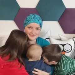 Mama w chustce i trójka dzieci. Najstarszy syn całuje maluszka, córka przytula się do mamy. W tle heksagony i poduszki. BIRADS 4a.