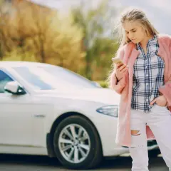 Młoda kobieta w różowym futrze i podartych dżinsach sprawdza telefon obok białego samochodu, być może po **szkodzie parkingowej**.