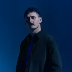 Dawid Podsiadło w ciemnej kurtce i fioletowej koszulce na niebieskim tle.