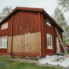 Ett rött trähus målas om. En stege står lutad mot huset, och färgburkar och penslar ligger utspridda på en presenning. Det ser ut som en perfekt dag att måla i solsken.