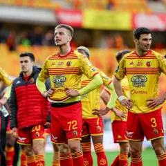 Zawodnicy Jagiellonii Białystok w żółto-czerwonych strojach stoją na boisku, gotowi do gry.