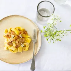 Ein Teller mit cremiger Pasta, die wie Fettuccine aussieht, garniert mit zarten Blüten und Speck. Daneben ein Glas Wasser und ein Strauß weißer Blumen.