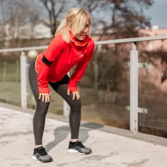 Kobieta w czerwonej bluzie i legginsach pochyla się, odpoczywając po treningu. Pokazuje, jak biegać, dbając o formę.