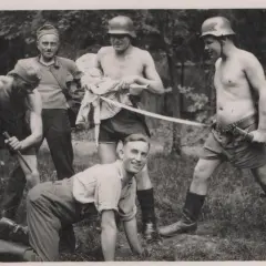 Fünf Männer, einige mit Helmen, posieren mit Waffen. Ein Mann hält eine Puppe. Die Szene erinnert an die Verbrechen der Wehrmacht.