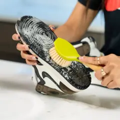 Main tenant une brosse verte, une personne nettoie la semelle extérieure d'une chaussure avec de la mousse.