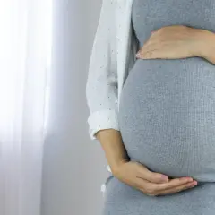 Zbliżenie na brzuch ciężarnej kobiety w szarej sukience, trzymający się za brzuch.