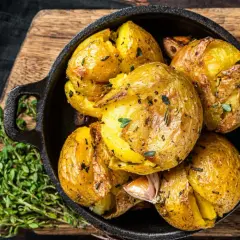Patatas al horno doradas y crujientes, con hierbas y ajo, servidas en una sartén de hierro fundido sobre una tabla de madera.