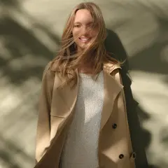 Uśmiechnięta blondynka w beżowym trenczu i swetrze. Idealne połączenie elegancji i komfortu, tworzące stylowe, sportowe stylizacje.
