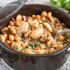 Ein Löffel voller cremiger dicke bohnen mit speck, garniert mit Petersilie, aus einem braunen Topf.