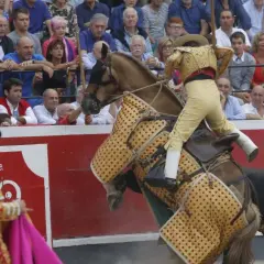 Un rejoneador, quizás Ana Romero, monta un caballo cubierto con un capote mientras un toro se acerca.
