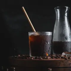 Ett glas med kallpressat kaffe, isbitar och en träpinne på en träbricka med kaffebönor. En karaff med kaffe står bredvid.
