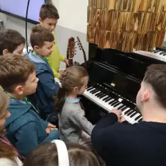 Dzieci słuchają lekcji muzyki przy pianinie. Nauczyciel pokazuje, do której klasy jest muzyka, a mali melomani z uwagą obserwują.