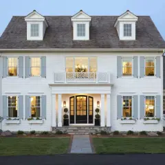 A stately white house with classic colonial architecture, featuring dormer windows, light blue shutters, and a welcoming porch.