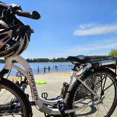 Rower z kaskiem czeka na plaży nad jeziorem, gdzie w upalny dzień kąpie się wiele osób. Idealne miejsce na wycieczkę rowerową i relaks w Puszczy Dulowskiej.
