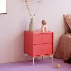 Ein Nachttisch in leuchtender **pink Farbe** steht neben einem Bett mit rosa Kissen. Dekorative Objekte und eine Vase mit Trockenblumen zieren die Oberfläche.