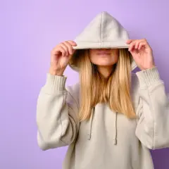 Kobieta w beżowej bluzie z kapturem, zasłaniająca twarz.
