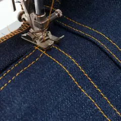 A sewing machine's topstitching foot creates neat, parallel yellow stitches on dark denim fabric, adding a professional finish.