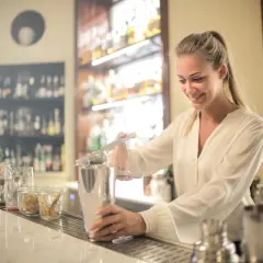 Uśmiechnięta barmanka przygotowuje drinka, ucząc się, jak zostać barmanem. Za nią półki z alkoholem.