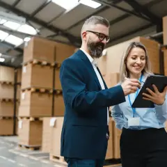 Uśmiechnięty logistyk i jego koleżanka analizują dane na tablecie w magazynie pełnym paczek.