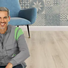 Un uomo sorridente in gilet da lavoro, pronto per il montaggio SPC. Sfondo con poltrona blu e parete decorata.