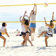 Grupa ludzi gra w siatkówkę plażową, odbijając piłkę nad siatką. Widać dynamiczne akcje i radość z gry, co oddaje zasady siatkówki plażowej.